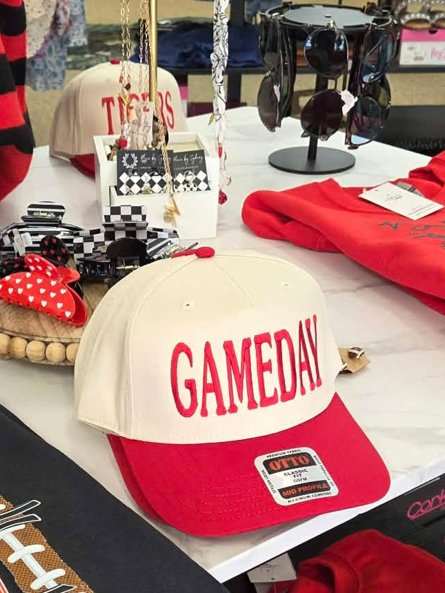 A retail display table showcasing various sports merchandise, including a black graphic t-shirt with football field and tiger graphics, a red graphic t-shirt, a red and cream baseball cap reading 'GAMEDAY', a red striped long-sleeve shirt, and other team apparel in a store setting.
